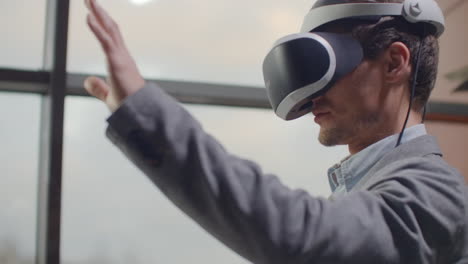 Mann-In-Einem-Helm-Der-Virtuellen-Realität-Vor-Dem-Hintergrund-Eines-Riesigen-Fensters-Im-Büro-Am-Arbeitsplatz.-Designingenieur,-Der-In-Einem-Virtual-Reality-Helm-Arbeitet.